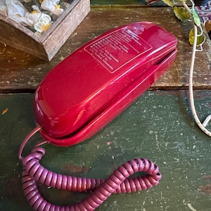 May include: A vintage red telephone with a coiled cord rests on a distressed green wooden surface. The phone has a glossy finish and a label with text. The coiled cord is a deep purple color.
