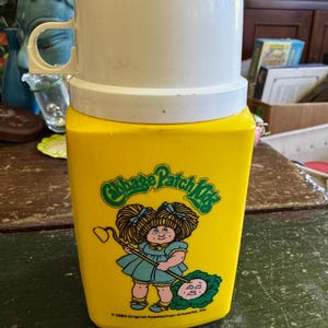 Op de afbeelding: Een gele plastic thermoskan met een witte schroefdop en handvat. De voorkant is voorzien van een Cabbage Patch Kids-afbeelding met een kind met bruin haar dat een schoffel vasthoudt naast een kool, met de tekst 1983 Original Appalachian Artworks, Inc.