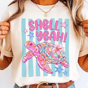 Könnte beinhalten: Cremefarbenes T-Shirt mit einem bunten Meeresschildkröten-Motiv und dem Text "SHELL YEAH!". Die Schildkröte ist rosa mit einer Blume und der Hintergrund hat blaue Streifen und Sterne.