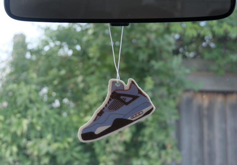 Puede incluir: Un ambientador con forma de zapatilla deportiva azul claro y marr&oacute;n colgando del espejo retrovisor de un coche. El ambientador, una r&eacute;plica de un dise&ntilde;o de zapatilla deportiva popular, cuelga de un cord&oacute;n blanco.