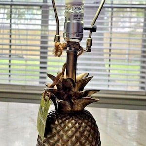 Puede incluir: Una lámpara de mesa dorada con forma de piña y superficie texturizada. La lámpara tiene una base de metal y una bombilla. La lámpara está sobre una mesa cerca de una ventana.