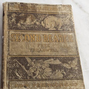 Puede incluir: Un libro antiguo "Second Reader" con una cubierta desgastada y texturizada. La cubierta presenta ilustraciones de una tortuga, un conejo y una figura con un bastón. El título y el nombre del autor están impresos en letras doradas. El libro es de Row, Peterson & Co.