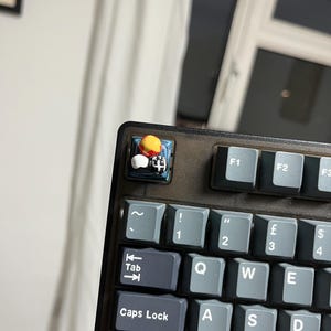 May include: Close-up of a mechanical keyboard with gray keycaps. The Esc key is replaced with a custom keycap featuring a small, colorful, 3D-printed design. The keyboard has a dark gray frame and is set against a blurred background.