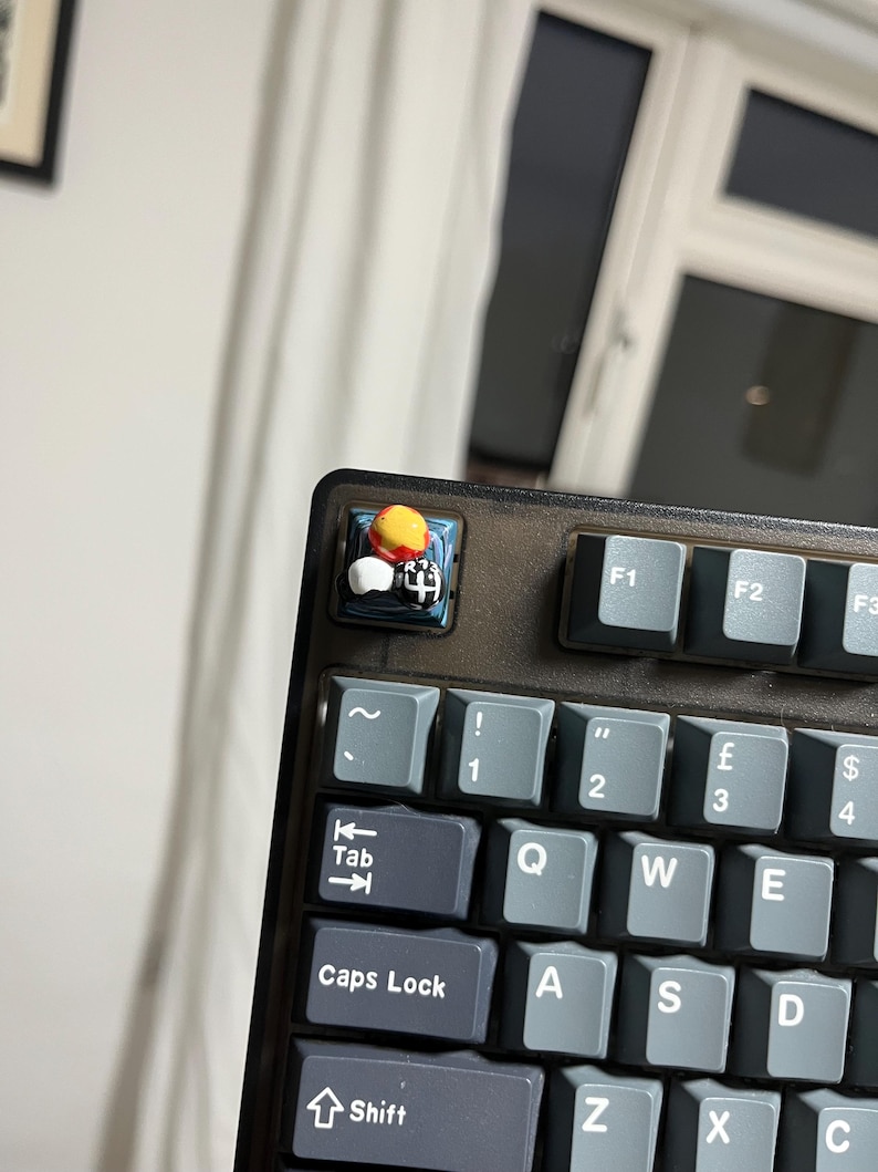 May include: Close-up of a mechanical keyboard with gray keycaps. The Esc key is replaced with a custom keycap featuring a small, colorful, 3D-printed design. The keyboard has a dark gray frame and is set against a blurred background.