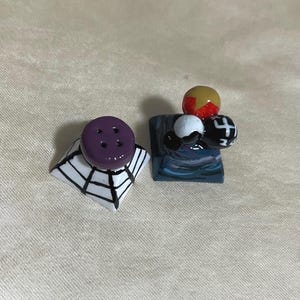 May include: Two decorative salt and pepper shakers. One shaker is purple with four holes and sits atop a white and black spiderweb design. The other shaker has a blue base with a cluster of colorful spheres.