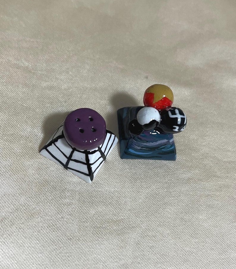 May include: Two decorative salt and pepper shakers. One shaker is purple with four holes and sits atop a white and black spiderweb design. The other shaker has a blue base with a cluster of colorful spheres.