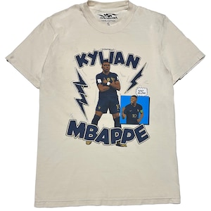 May include: Beige cotton t-shirt with a graphic of Kylian Mbapp&eacute; in a blue football kit. The design includes the name "KYLIAN MBAPPE" in navy blue, lightning bolt accents, and a small image of the player. The shirt has a crew neck and short sleeves.