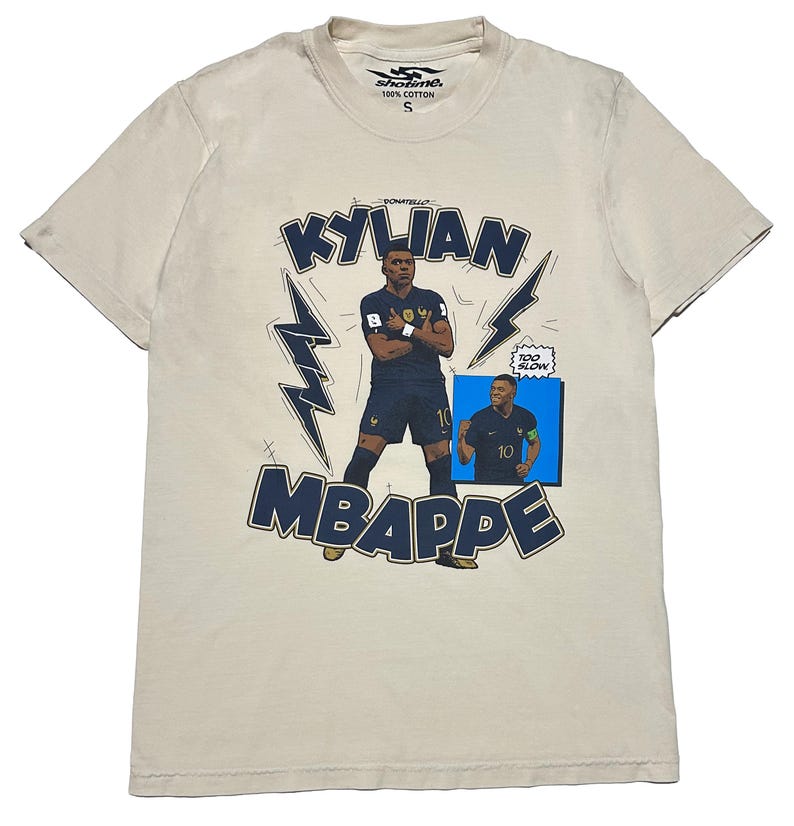 May include: Beige cotton t-shirt with a graphic of Kylian Mbapp&eacute; in a blue football kit. The design includes the name "KYLIAN MBAPPE" in navy blue, lightning bolt accents, and a small image of the player. The shirt has a crew neck and short sleeves.