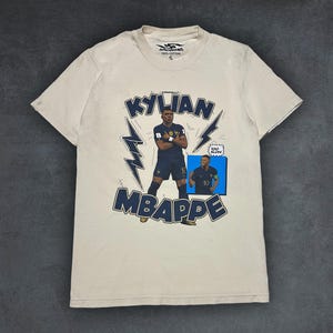 May include: Cream-coloured t-shirt with a navy blue graphic of Kylian Mbapp&eacute; in a French football kit. The design includes the name "KYLIAN MBAPPE", lightning bolt accents, and a small image of the player. The shirt is made of 100% cotton.