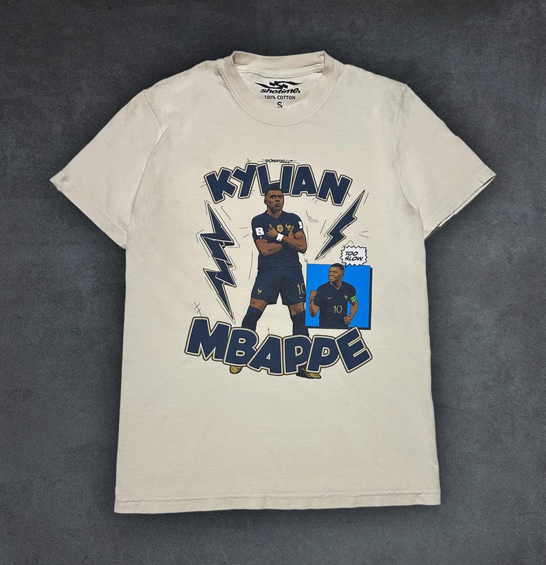 May include: Cream-coloured t-shirt with a navy blue graphic of Kylian Mbapp&eacute; in a French football kit. The design includes the name "KYLIAN MBAPPE", lightning bolt accents, and a small image of the player. The shirt is made of 100% cotton.