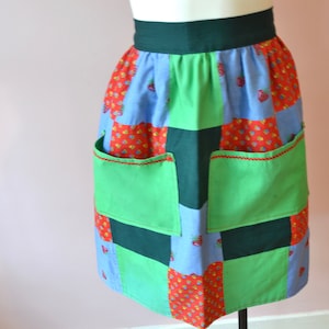 以下が含まれることがあります： 緑色のウエストバンドとポケットが付いたパッチワークエプロン。エプロンは赤、青、緑の生地の正方形が特徴です。赤い正方形には小さなイチゴ模様があります。ポケットは赤いステッチでトリミングされています。