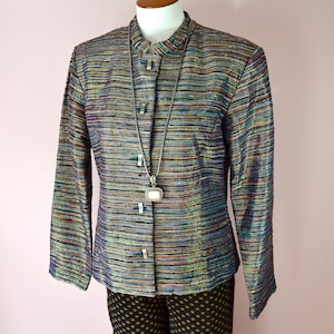 May include: A multi-colored, horizontally striped jacket with a mandarin collar and long sleeves. The jacket has three rectangular buttons and is paired with black patterned pants. A silver necklace with a square pendant hangs from the jacket.