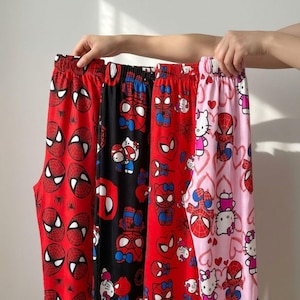 May include: Four pairs of pajama pants with elastic waistbands are displayed. The pants feature various patterns, including Spider-Man faces, Spider-Man with hearts, and a combination of Spider-Man and Hello Kitty characters. The color palette includes red, black, and pink.