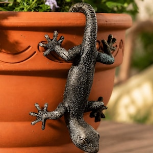 May include: A dark bronze-colored lizard figurine with a textured, scaly surface and a long, curved tail. The lizard is positioned vertically, clinging to the side of a large, round, terracotta-colored clay plant pot with a smooth finish.