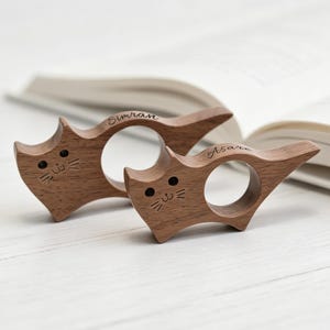 Puede incluir: Dos sujetadores de páginas de madera marrón con forma de gato y orificios circulares para los dedos. Cada pieza presenta detalles faciales grabados, incluidos ojos y bigotes, con los nombres Simran y Asare grabados en los bordes superiores.