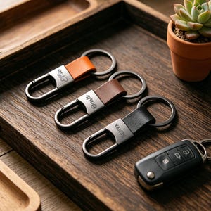 May include: Three leather keyrings in orange, brown, and black, each with a silver clasp and a name engraved on the leather. A black car key fob is also present. The items are arranged on a wooden tray.