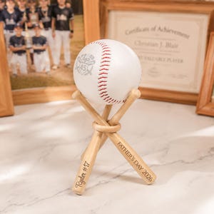 Może przedstawiać: Biała piłka baseballowa z czerwonymi szwami, ozdobiona szarą grafiką przedstawiającą dwie skrzyżowane pałki oraz napis PROUD DAD 45. Spoczywa na stojaku z trzech miniaturowych drewnianych pałek baseballowych z wygrawerowanym napisem Ryder #17 i FATHER'S DAY 2026.