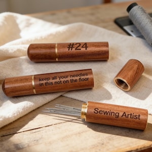 Puede incluir: Estuches de agujas de madera con detalles de latón. Un estuche está abierto, revelando agujas. Los estuches tienen texto que dice "#24", "keep all your needles in this not on the floor" y "Sewing Artist". Sobre una superficie de madera.