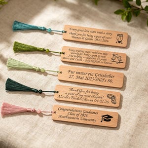 Puede incluir: Marcadores de libros de madera con borlas de colores. Cada marcador tiene un mensaje y un diseño únicos, que incluyen un birrete, anillos de boda y copas de champán. Las borlas son en tonos de verde azulado, verde y rosa.
