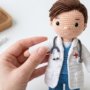 May include: A crocheted doctor doll wearing a white coat with a red cross, blue scrubs, and a gray stethoscope. The doll has brown hair and is being held by a hand.