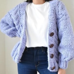 Op de afbeelding: Een lichtpaars, grofgebreid vest met grote, donkerbruine knopen. De open trui heeft een kabelgebreid ontwerp op de mouwen en voorpanelen. Het vest wordt gedragen over een wit t-shirt en een donkerblauwe jeans.