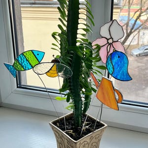 以下が含まれることがあります： 色とりどりのステンドグラスのガーデンピックが付いた鉢植えのサボテン。青と緑の魚、黄と白の蝶、ピンクと白の花、青い鳥、オレンジ色のじょうろが含まれています。鉢は正方形でベージュ色、装飾的な模様があります。