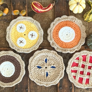 Peut inclure: Cinq dessous de verre ronds au crochet conçus pour ressembler à diverses tartes, avec des bords beiges festonnés. L'ensemble comprend une tarte à la banane jaune, une tarte à la citrouille orange, une tarte au chocolat marron et une tarte rouge.