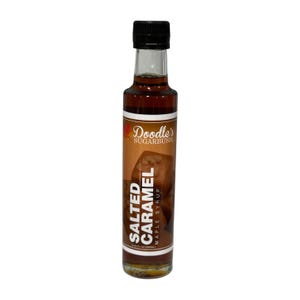May include: A clear glass bottle of Doodle's Sugarbush Salted Caramel Maple Syrup. The bottle has a black cap and a brown label with the product name and a caramel image. The bottle contains a dark amber liquid.