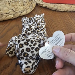 May include: A plush leopard-print toy with black eyes and a black mustache. The toy is lying on a wooden surface. A heart-shaped tag is attached, with the text "Freckles" and "style 4066".