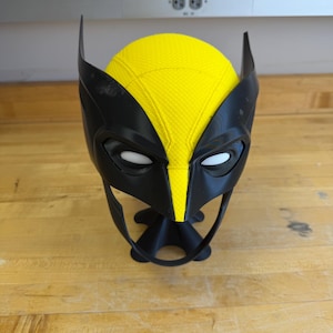 May include: A black and yellow Wolverine mask with white eye openings. The mask has pointed ears and a yellow centre section. The mask is resting on a wooden surface.