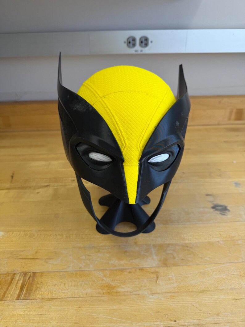 May include: A black and yellow Wolverine mask with white eye openings. The mask has pointed ears and a yellow centre section. The mask is resting on a wooden surface.