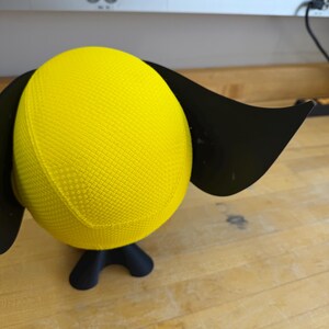 May include: A yellow, textured sphere with black, wing-like attachments and a black base. The sphere is a bright yellow colour. The base has three curved legs. The object is on a wooden surface.