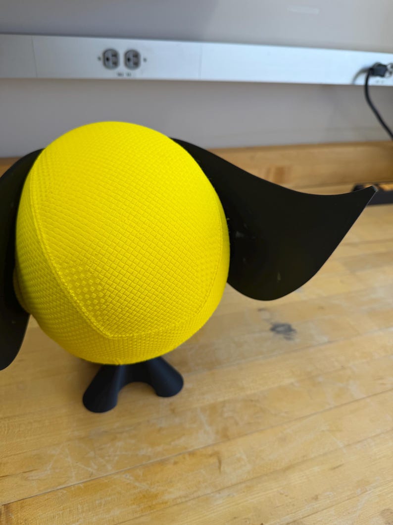 May include: A yellow, textured sphere with black, wing-like attachments and a black base. The sphere is a bright yellow colour. The base has three curved legs. The object is on a wooden surface.
