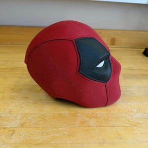 May include: A red Deadpool mask with black eye and mouth details. The mask is made of a textured fabric and is designed to cover the entire head. The mask is resting on a wooden surface.