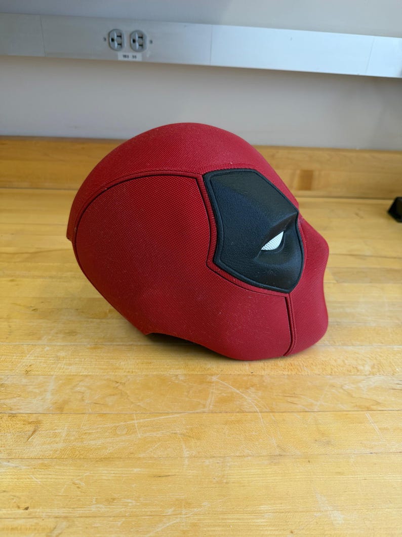 May include: A red Deadpool mask with black eye and mouth details. The mask is made of a textured fabric and is designed to cover the entire head. The mask is resting on a wooden surface.