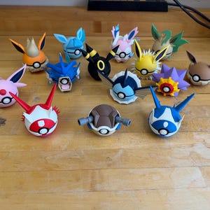 Może przedstawiać: Kolekcja kolorowych, stylizowanych figurek różnych postaci Pokémon. Figurki są okrągłe z detalami, w tym uszami, rogami i innymi charakterystycznymi cechami. Paleta kolorów obejmuje pomarańczowy, niebieski, różowy, żółty, czarny i brązowy.