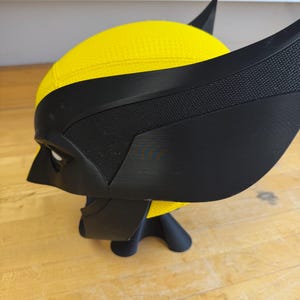 May include: A yellow and black Wolverine mask, with the character's distinctive pointed ears and eye openings. The mask is made of a textured yellow material and black accents, and is displayed on a black stand.
