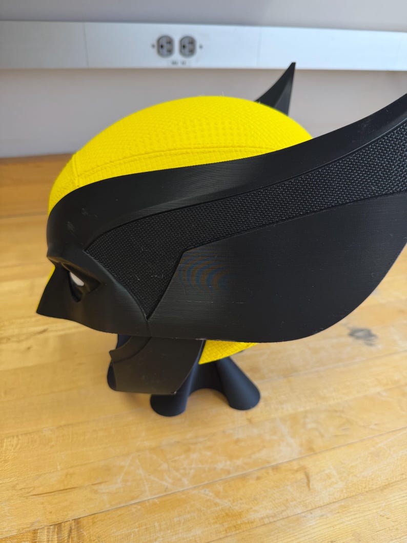 May include: A yellow and black Wolverine mask, with the character's distinctive pointed ears and eye openings. The mask is made of a textured yellow material and black accents, and is displayed on a black stand.