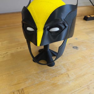 May include: A black and yellow Wolverine mask, with a yellow top and black eye and ear details. The mask is displayed on a black stand. The mask is made of plastic and is likely a cosplay or costume accessory.