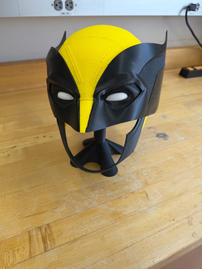 May include: A black and yellow Wolverine mask, with a yellow top and black eye and ear details. The mask is displayed on a black stand. The mask is made of plastic and is likely a cosplay or costume accessory.