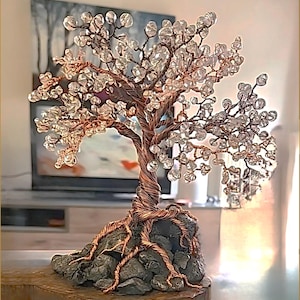 Könnte beinhalten: Eine dekorative Baumskulptur aus gedrehtem Kupferdraht mit klaren, facettierten Kristallperlen an den Zweigen. Der Baum ist auf einer Basis aus dunklen, strukturierten grauen Steinen verankert, wobei Kupferdrahtwurzeln darüber verlaufen.
