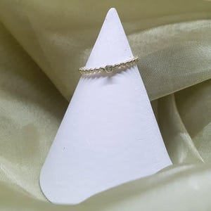 Peut inclure: Une délicate bague en or avec une petite pierre précieuse transparente au centre. La bague est ornée de minuscules perles espacées régulièrement. Elle est présentée sur un support blanc en forme de cône, sur un fond de tissu vert clair.