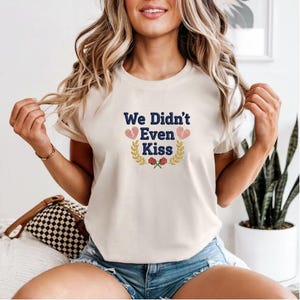 Op de afbeelding: Crèmekleurig T-shirt met de geborduurde tekst "We Didn't Even Kiss" in marineblauw. Het ontwerp bevat gebroken harten, een lauwerkrans en rode rozen. Het shirt is een casual stijl met korte mouwen.