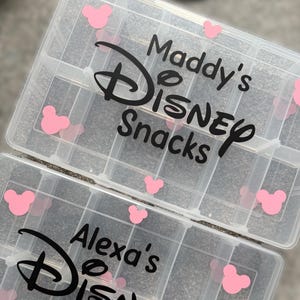 May include: Two clear plastic snack organizers with multiple compartments. Each organizer has the words "Maddy's Disney Snacks" and "Alexa's Disney Snacks" in black script, along with pink Mickey Mouse head accents.