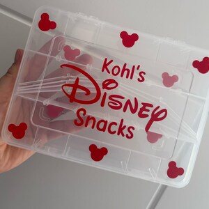 May include: A clear plastic storage container with multiple compartments. The container has red Mickey Mouse head shapes and the words "Kohl's DISNEY Snacks" in red lettering. The container is held by a person.