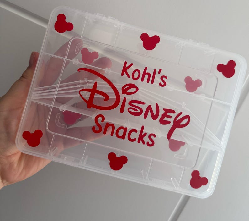 May include: A clear plastic storage container with multiple compartments. The container has red Mickey Mouse head shapes and the words "Kohl's DISNEY Snacks" in red lettering. The container is held by a person.