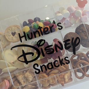 May include: A clear plastic snack box with multiple compartments filled with various treats. The box is labeled "Hunter's Disney Snacks" in black script. Snacks include cookies, candies, pretzels, and marshmallows. A Mickey Mouse head is visible in the top right corner.