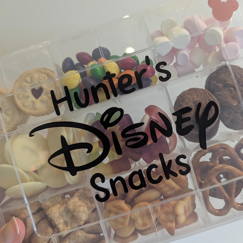 May include: A clear plastic snack box with multiple compartments filled with various treats. The box is labeled "Hunter's Disney Snacks" in black script. Snacks include cookies, candies, pretzels, and marshmallows. A Mickey Mouse head is visible in the top right corner.