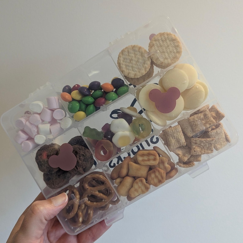 May include: A clear plastic container filled with various snacks, including crackers, candies, pretzels, and marshmallows. The snacks are arranged in separate compartments. A hand is holding the container.