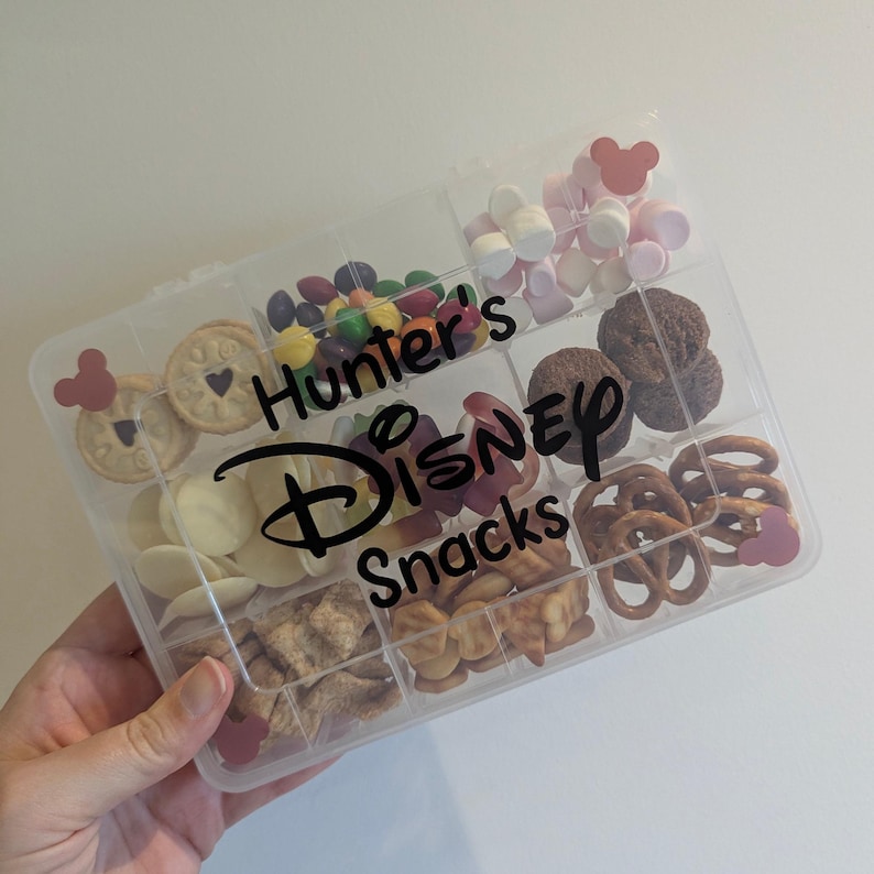 May include: A clear plastic snack box filled with various treats, including cookies, candies, pretzels, and marshmallows. The box is divided into compartments and features the text "Hunter's Disney Snacks" in black lettering, with Mickey Mouse head accents.